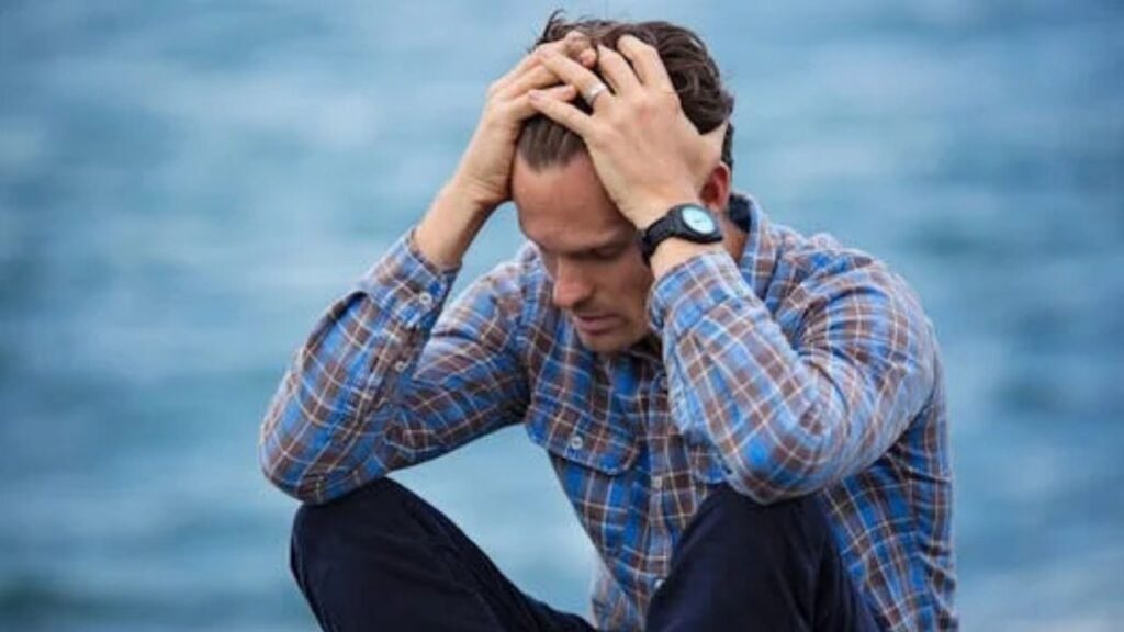 A man in a plaid shirt sits by the water looking distressed, symbolizing stress