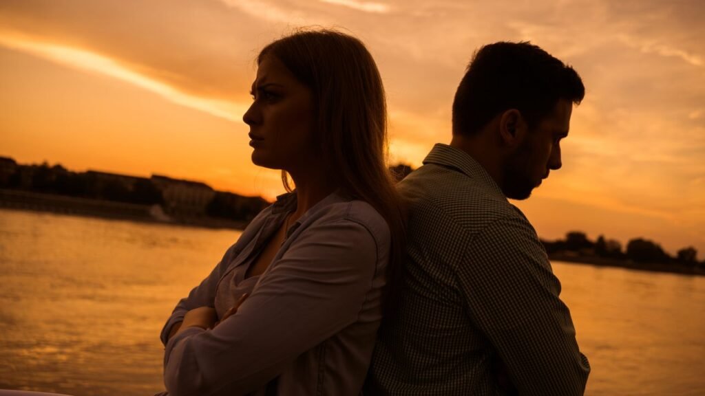 Man and woman standing back to back at sunset near water, showing emotional distance