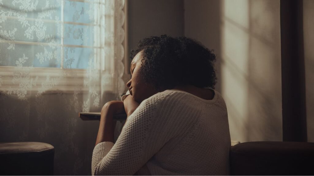 Person sitting by a window with head resting on arms, looking thoughtful in soft natural light