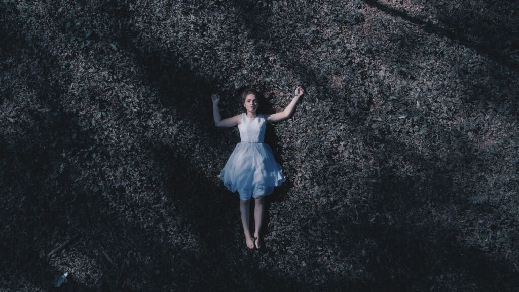 Woman lying on forest ground with arms open, wearing a light dress in a moody setting