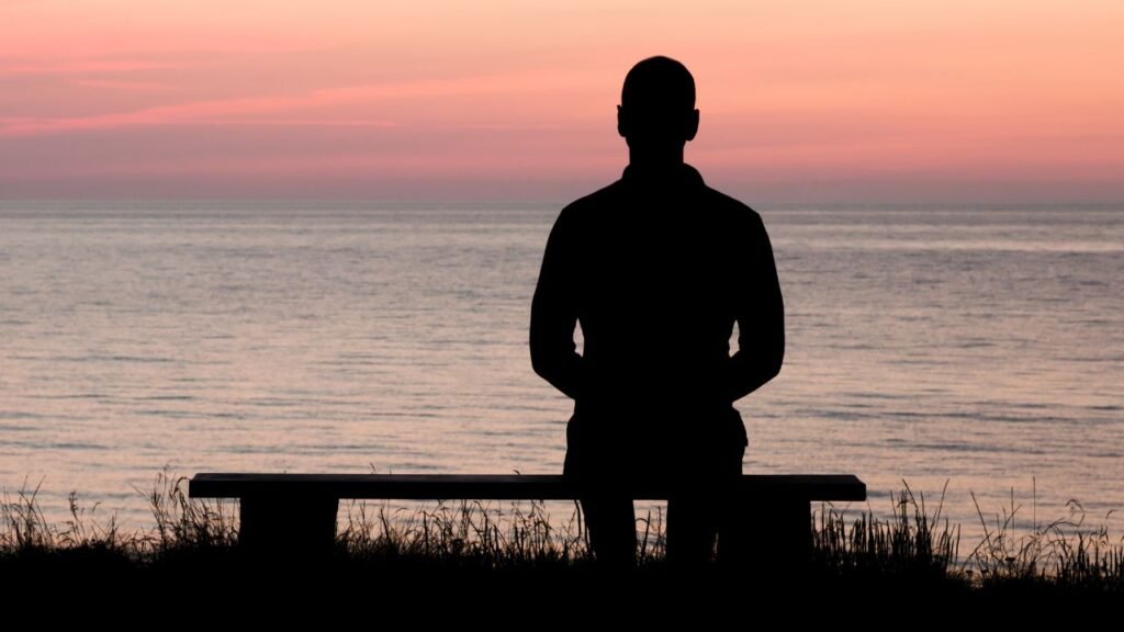 Photograph of a person sitting on a bench facing a calm ocean during sunset, with a pink and orange sky. Silhouette of the person and bench contrasts with soft colors of water and sky, evoking a peaceful and contemplative mood.