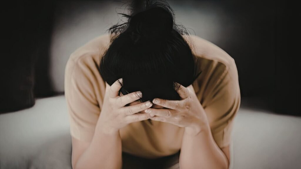 Photograph of a person sitting on a couch with head bowed and hands covering face, conveying feelings of stress or distress. The person wears a light brown shirt and has dark hair tied up, with a blurred dark background emphasizing emotional focus.