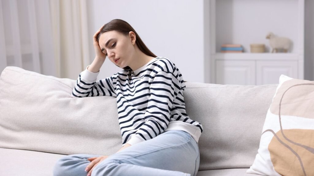 Photograph of a person sitting on a light-colored couch in a bright room, wearing a black and white striped long-sleeve shirt and light blue jeans. The person is resting their head on one hand, suggesting a contemplative or tired mood, with a white pillow featuring abstract brown and white patterns nearby.