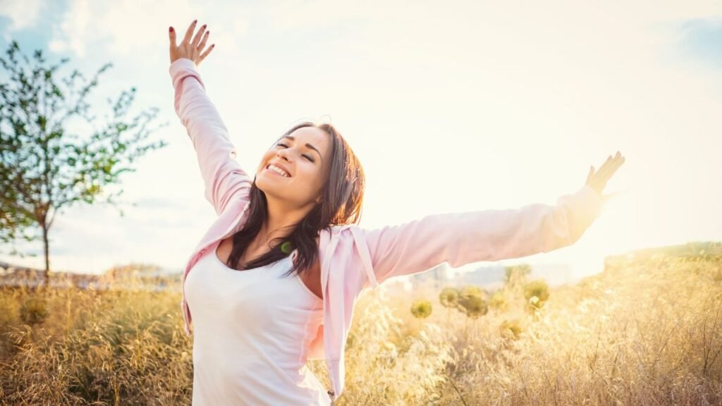7 Signs You’re Becoming Your Real Self During Identity Shift 12 Photograph of a person standing outdoors in a field with arms raised, expressing joy or freedom. The scene features natural sunlight, tall grasses, and a tree in the background, highlighting a serene and uplifting moment.