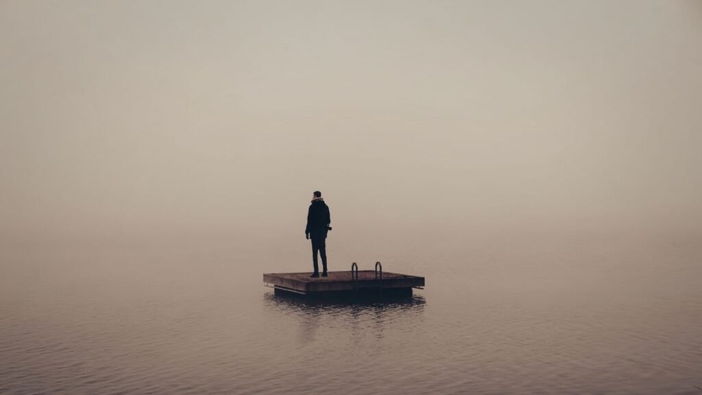 Photograph of a solitary person standing on a small wooden platform floating on calm water, surrounded by dense fog. The muted color palette and minimal composition evoke a sense of isolation and tranquility.