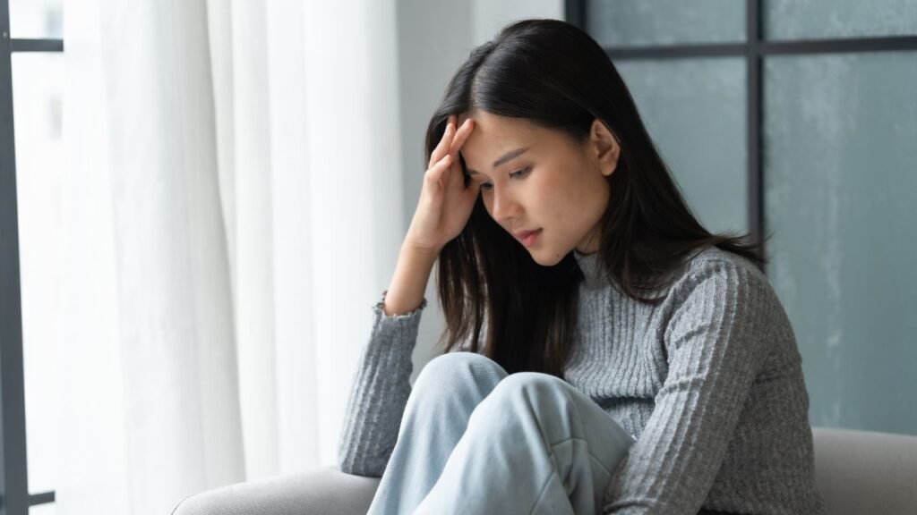Photograph of a woman sitting on a couch indoors, wearing a gray ribbed sweater and light blue pants. She is resting her head on her hand, suggesting a contemplative or tired mood, with natural light coming through a window in the background.