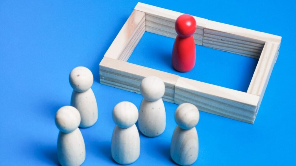 Photograph showing five white wooden figurines grouped together outside a rectangular wooden frame containing a single red figurine. The composition visually represents concepts of isolation, exclusion, or separation using contrasting colors and spatial arrangement.