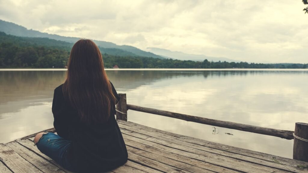 a person sitting cross-legged on a wooden dock facing a calm lake with mountains and a cloudy sky in the background. The scene conveys tranquility and reflection, with muted colors and natural elements emphasizing peaceful solitude.