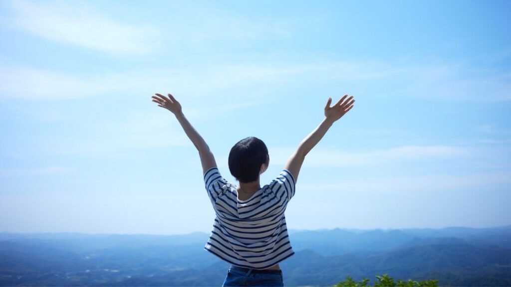 A person standing on a mountain peak with arms raised, facing a vast landscape of rolling hills and a clear blue sky. The scene conveys a sense of freedom, achievement, and connection with nature.