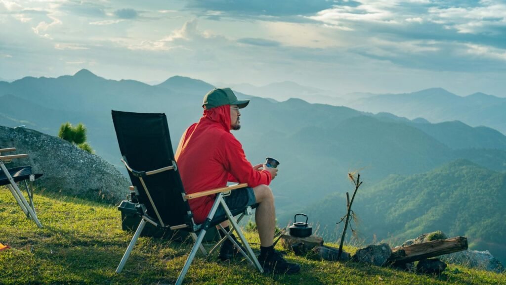 7 Clear Signs of Personal Growth Even if You Don’t Feel Like Growing 4 A person wearing a red jacket and green cap sitting on a folding chair on a grassy hilltop, holding a cup and looking at a mountainous landscape under a cloudy sky. The scene includes camping gear such as a kettle and logs near a small fire pit, highlighting a peaceful outdoor camping moment